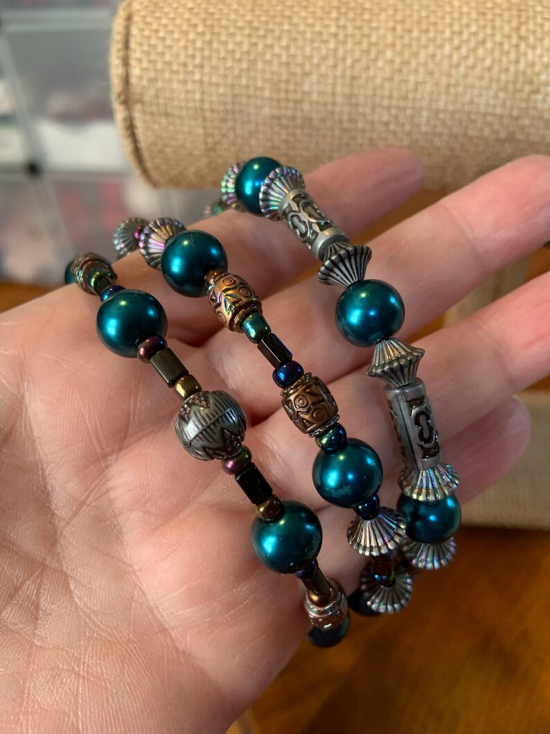 May include: Three teal beaded bracelets with decorative metal elements. The bracelets feature a mix of round and cylindrical beads, with silver and copper-toned metal accents. The bracelets are designed to wrap around the wrist.