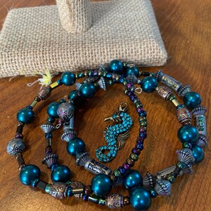 May include: A set of beaded bracelets and a seahorse charm. The bracelets have teal beads, metallic details, and decorative components. The seahorse charm is embellished with blue gemstones.