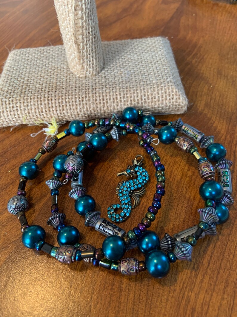 May include: A set of beaded bracelets and a seahorse charm. The bracelets have teal beads, metallic details, and decorative components. The seahorse charm is embellished with blue gemstones.