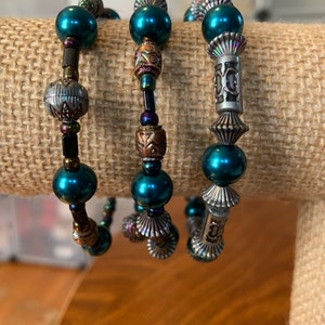 May include: Three beaded bracelets with teal faux pearls and decorative metal beads. The bracelets feature silver and bronze metal accents, creating a layered look. The bracelets are displayed on a neutral-toned surface. The beads are measured in centimetres.