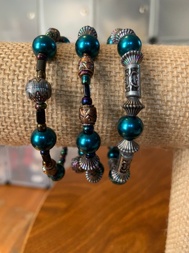 May include: Three beaded bracelets with teal faux pearls and decorative metal beads. The bracelets feature silver and bronze metal accents, creating a layered look. The bracelets are displayed on a neutral-toned surface. The beads are measured in centimetres.