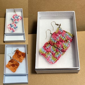 May include: Three pairs of crocheted earrings in a white box. The earrings are in different colours and patterns. The first pair is blue and white, the second pair is pink, green, and blue, and the third pair is orange.