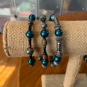 May include: Three beaded bracelets with teal beads and decorative metal accents. The bracelets are displayed on a tan fabric surface. The beads are interspersed with ornate metal spacers and charms, creating a layered look.