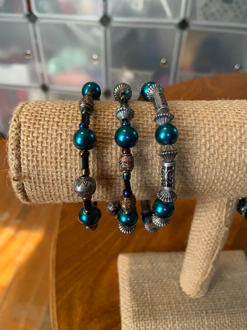 May include: Three beaded bracelets with teal beads and decorative metal accents. The bracelets are displayed on a tan fabric surface. The beads are interspersed with ornate metal spacers and charms, creating a layered look.