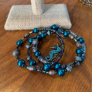 May include: Three beaded bracelets with teal faux pearls and decorative metal elements. One bracelet features a seahorse charm with blue rhinestones. The bracelets are displayed on a wooden surface.