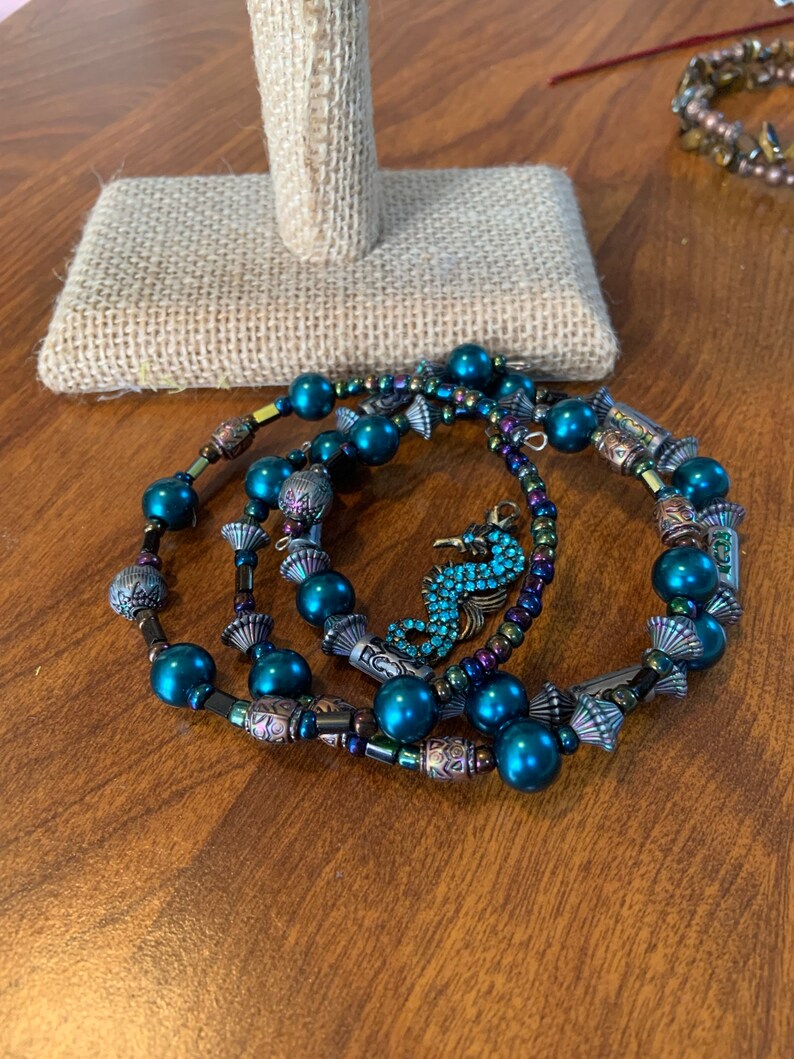 May include: Three beaded bracelets with teal faux pearls and decorative metal elements. One bracelet features a seahorse charm with blue rhinestones. The bracelets are displayed on a wooden surface.