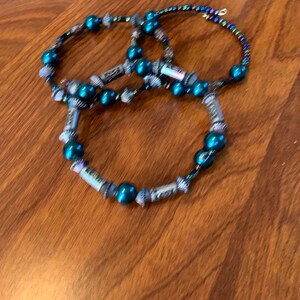 May include: Three beaded bracelets. Each bracelet features teal beads and silver decorative elements. One bracelet has iridescent beads. The bracelets are arranged on a wooden surface.