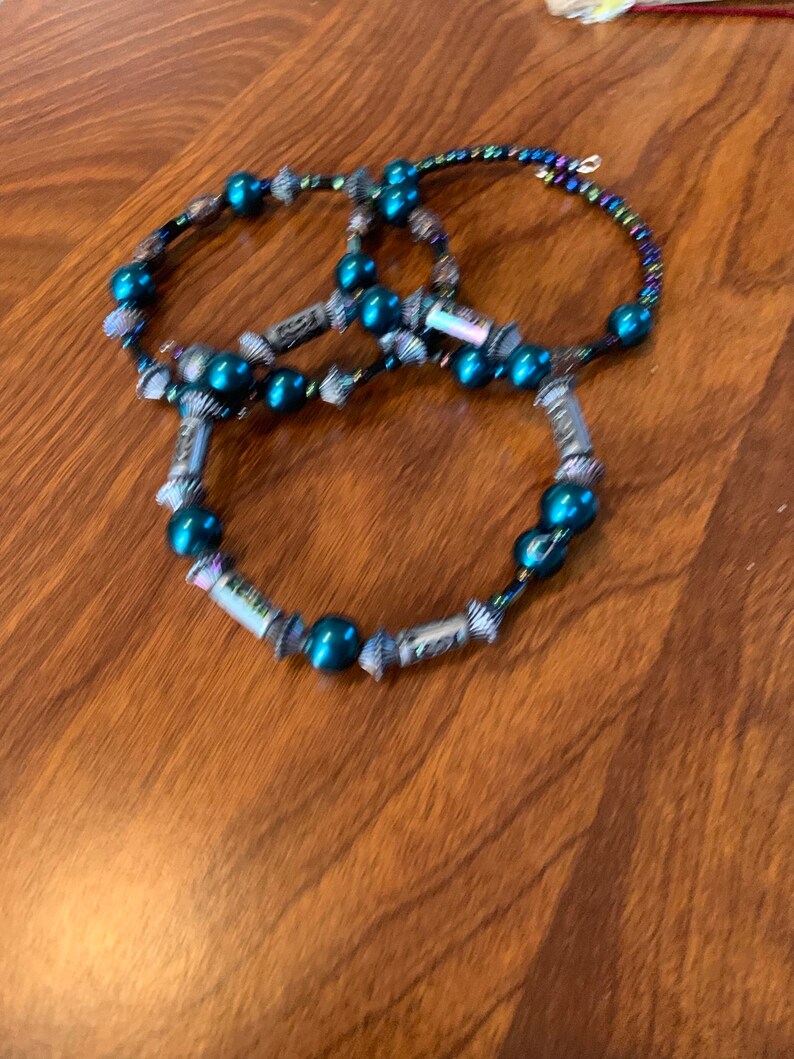 May include: Three beaded bracelets. Each bracelet features teal beads and silver decorative elements. One bracelet has iridescent beads. The bracelets are arranged on a wooden surface.