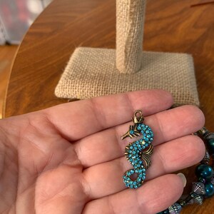 May include: A bronze seahorse charm, adorned with turquoise blue crystals. The charm is held in a hand, with a beaded bracelet in the background. The seahorse design features detailed craftsmanship.