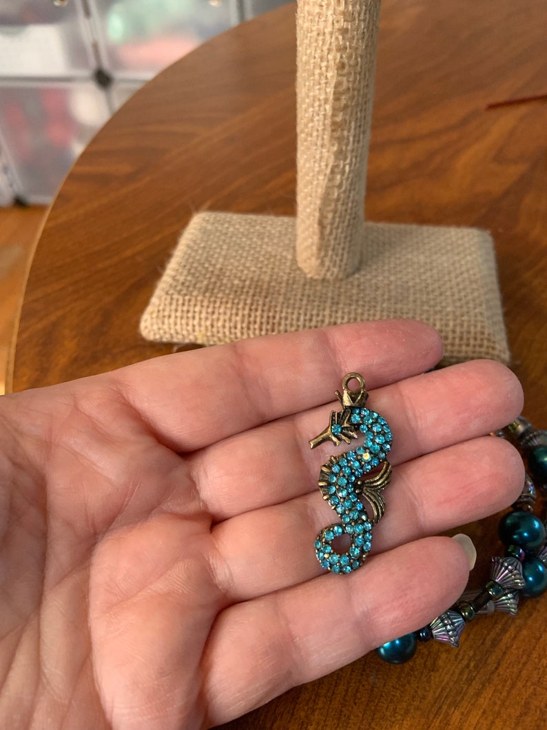 May include: A bronze seahorse charm, adorned with turquoise blue crystals. The charm is held in a hand, with a beaded bracelet in the background. The seahorse design features detailed craftsmanship.