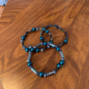 May include: Three teal beaded bracelets with silver accents are displayed on a wooden surface. The bracelets feature dark teal beads and decorative silver elements. The beads are strung on a dark cord, creating a layered effect.