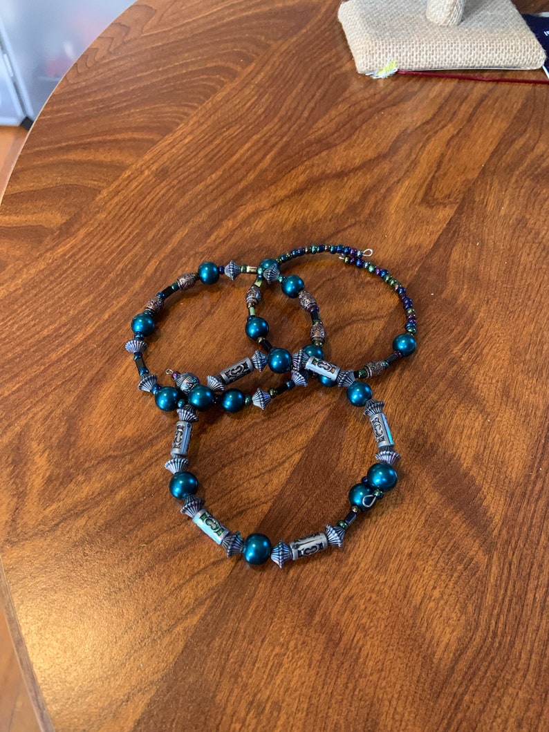 May include: Three teal beaded bracelets with silver accents are displayed on a wooden surface. The bracelets feature dark teal beads and decorative silver elements. The beads are strung on a dark cord, creating a layered effect.