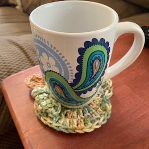 May include: A white ceramic mug with a blue, green and yellow paisley design. The mug is sitting on a crocheted coaster with green, brown and yellow yarn.