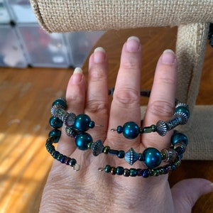 May include: A teal bead and iridescent accent spiral wrap bracelet. The bracelet has multiple strands of beads, including large teal orbs and smaller, iridescent beads. Decorative metal elements add detail.