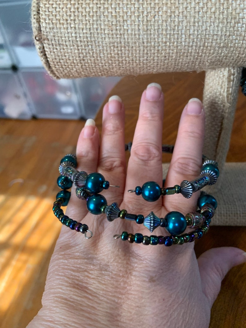 May include: A teal bead and iridescent accent spiral wrap bracelet. The bracelet has multiple strands of beads, including large teal orbs and smaller, iridescent beads. Decorative metal elements add detail.