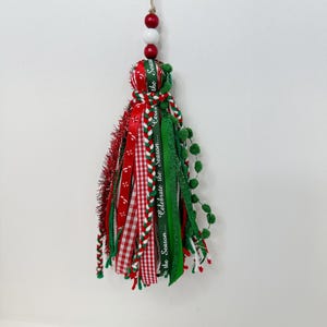 May include: A festive Christmas tassel ornament. The ornament features red, green, and white ribbons, including a gingham pattern and ribbons with the text "Celebrate the Season...". It is topped with red and white beads and a twine loop.