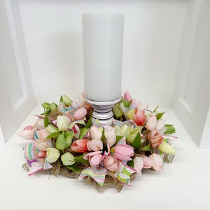 May include: A white candle holder with a pink and white tulip wreath. The wreath is made of burlap and ribbon.