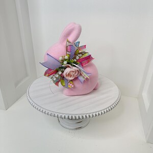 May include: A pink bunny figurine with a decorative bow and floral accents. The bunny is seated on a white, distressed-look cake stand. The bow features colorful ribbons and small flowers, adding a festive touch to the Easter-themed decor.