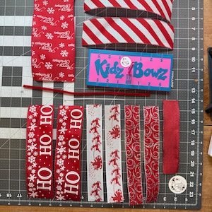 May include: A collection of craft supplies including red ribbon with Merry Christmas text, red and white striped ribbon, a blue Kidz Bowz tool, a red pipe cleaner, a white zip tie, red ribbon with OH HO HO text, and white ribbon with red reindeer patterns.