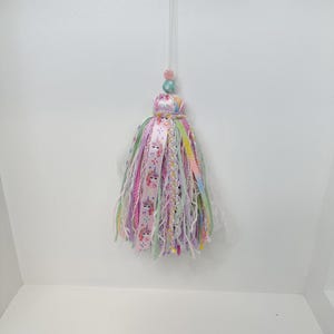 May include: A colorful tassel ornament with a clear hanging string. The tassel features ribbons and fabric in shades of pink, purple, green, and yellow. Some ribbons have unicorn designs. The top has decorative beads.
