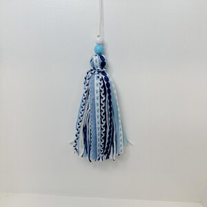 May include: A decorative tassel in shades of blue and white, hanging against a white background. The tassel features a mix of patterned and solid ribbons, with a light blue bead and a white bead at the top. The tassel is suspended by a white string.