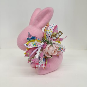 May include: Pink felt Easter bunny with a large bow made of colorful ribbons, floral accents, and a pink flower.