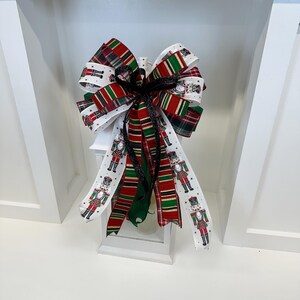 May include: A decorative Christmas bow featuring a mix of ribbons in red, green, and white. Some ribbons have a plaid pattern, while others display a nutcracker soldier design. The bow is tied with a black ribbon and is placed on a white lantern.