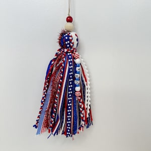 May include: A patriotic tassel with red, white, and blue ribbons and beads. The tassel features the letters "USA" on white and blue beads. The top has a wooden bead and a red bead, and is suspended by a thin string.