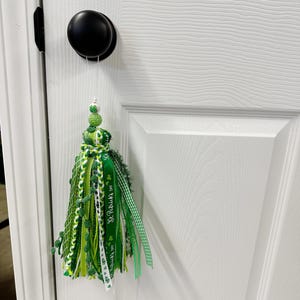 May include: A St. Patrick's Day tassel decoration hanging from a black doorknob. The tassel is made of various shades of green ribbons, mesh, and beads. Some ribbons have "St. Patrick's Day" printed on them, and others have shamrock patterns.