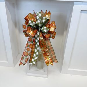 May include: A decorative fall bow featuring ribbons with pumpkin, leaf, and plaid patterns. The bow includes orange, green, and white colors. It is displayed on a white surface, suggesting a seasonal home decor item.