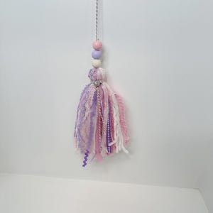 May include: A handmade tassel ornament with a string of pink, purple, and white beads. The tassel is made of various fabrics in shades of pink, purple, and white, with a silver charm at the top. The ornament hangs from a silver string.