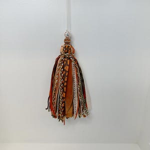 May include: A decorative tassel with a clear hanging loop. The tassel features a mix of ribbons in shades of orange, brown, and cream, with patterned and textured details. A beaded section with a small floral design sits above the ribbons.