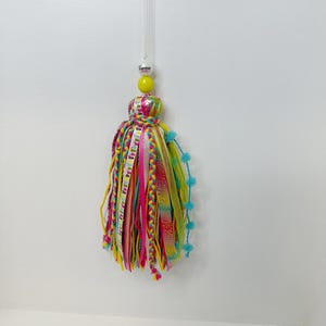 May include: A colorful tassel ornament with a yellow bead and a clear hanging string. The tassel features ribbons in various colors, including pink, yellow, and green. The ornament has a small white tag that says "Summer Vibes".