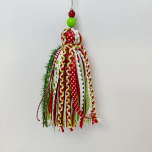May include: A festive Christmas tassel ornament. The ornament features a red, white, and green color scheme with a variety of ribbons, including polka dots and gingham patterns. The top has red and green beads.