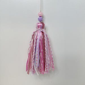 May include: A decorative tassel in shades of pink and purple, suspended by a clear string. The tassel features various ribbons, including some with the text "IT'S A GIRL". The top has a cluster of beads in pink and purple.