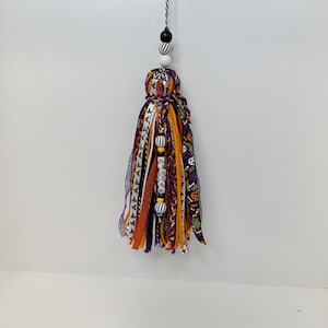 May include: A Halloween-themed tassel with orange, purple, and black ribbons and fabric strips. The tassel is adorned with beads and the word "Spooky" on a ribbon. It hangs from a black and white string.