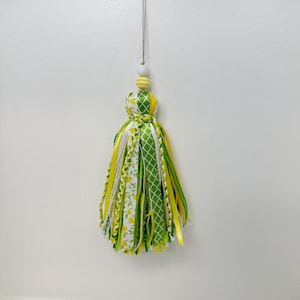 May include: A decorative tassel with a white wooden bead and a yellow and white striped bead at the top. The tassel is made of ribbons in various patterns, including lemons, green lattice, and solid yellow and white. It hangs from a thin, brown string.