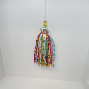 May include: A colorful tassel ornament with a yellow bead and a clear hanging string. The tassel features ribbons in various colors, including pink, blue, yellow, and green, with floral and zigzag patterns. Small blue pom-poms are attached to the side.