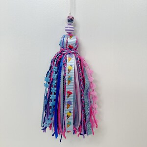 May include: A colorful tassel with a variety of ribbons and yarn in shades of pink, blue, and purple. The tassel is adorned with a ribbon featuring ice cream cones and is topped with striped beads and a small decorative bead.