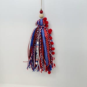 May include: A patriotic tassel ornament with red, white, and blue ribbons, braids, and pom-poms. The tassel has a wooden bead and a red bead at the top, with the word "AMERICA" spelled out on a white strand. This decorative item is perfect for adding a festive touch.