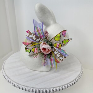 May include: A white bunny figurine with a colorful bow featuring Easter egg and bunny patterns. The bow includes pink, green, and purple ribbons, and a small pink rose. The bunny is displayed on a white, beaded cake stand.