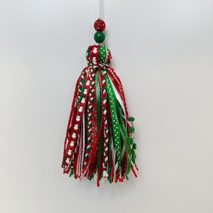 May include: A festive Christmas tassel ornament. The ornament features red, green, and white ribbons with snowman and polka dot patterns. A red beaded sphere and a green bead are at the top, with a clear string for hanging.