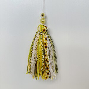 May include: A decorative tassel with a sunflower theme. The tassel features yellow, green, and white ribbons, lace, and fabric strips. It is adorned with a sunflower print and pearl accents. The tassel is suspended by a clear string and topped with yellow and white beads.