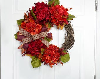 May include: A fall-themed wreath with vibrant red and orange hydrangeas, accented by green leaves and a patterned ribbon. The wreath is made of woven twigs and hangs on a white door.