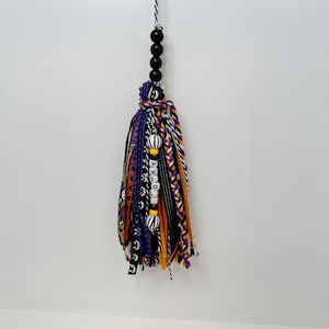 May include: A Halloween-themed tassel with black, purple, orange, and white accents. The tassel is made of various ribbons and yarn, with the word "Spooky" spelled out in white beads. Black wooden beads and a black and white cord complete the design.