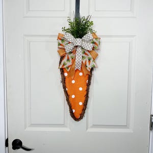 May include: A carrot-shaped Easter wreath with an orange fabric base and white polka dots. The wreath is trimmed with a brown woven border and topped with a green leafy accent and a decorative bow with carrot and polka dot patterns.