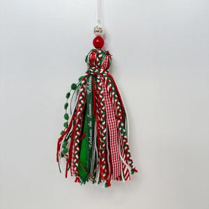 May include: A festive Christmas tassel ornament. The ornament features red, green, and white ribbons, with a red and white gingham pattern. A green ribbon with the text "Celebrate the Season" is also included. The ornament is topped with a red bead and a white bead with the text "I love Santa".