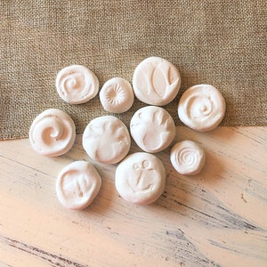 May include: A collection of small, white, round, decorative stones with various embossed designs, including swirls, a leaf, and an anchor. The stones are arranged on a distressed, light-colored wooden surface with a burlap background.