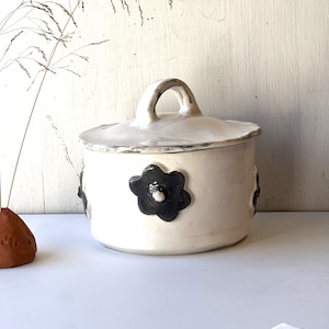 Peut inclure: Un pot en céramique blanche avec un couvercle et une poignée. Le pot est décoré d'un motif floral noir et blanc.