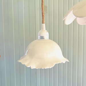 Puede incluir: Una lámpara colgante de cerámica blanca con un borde ondulado en forma de flor. La lámpara está suspendida por un cable de tela trenzada de color tostado. Una pequeña etiqueta blanca en la base muestra el texto 50Hz y MAX 40W de potencia.